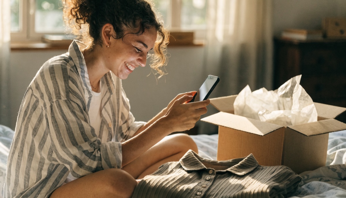 Mulher sentada na cama fotografando uma blusa com o celular ao lado de uma encomenda aberta