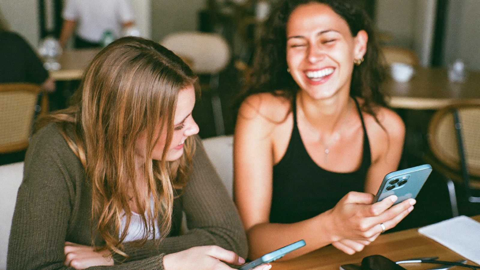Dois amigos sentados à mesa de um café comparando seus celulares e rindo