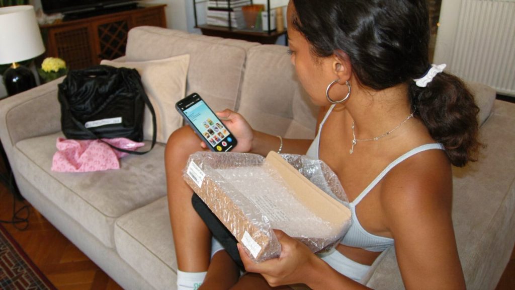 Mulher sentada no sofá olhando o celular com uma encomenda aberta no colo